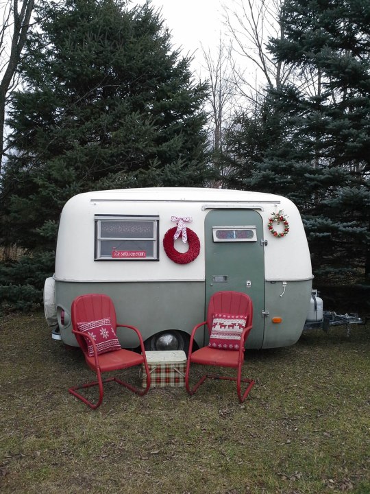 Green and white Boler travel trailer decorated for Christmas, with 2 red chairs in front, a vintage, plaid cooler, red wreath, bell wreath and "Santa, I can explain" sign.